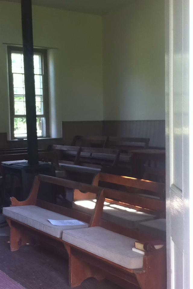 Photo looking into the Meetinghouse, showing Meetinghouse benches (wooden, with cushions) in foreground, wood stove and window in background