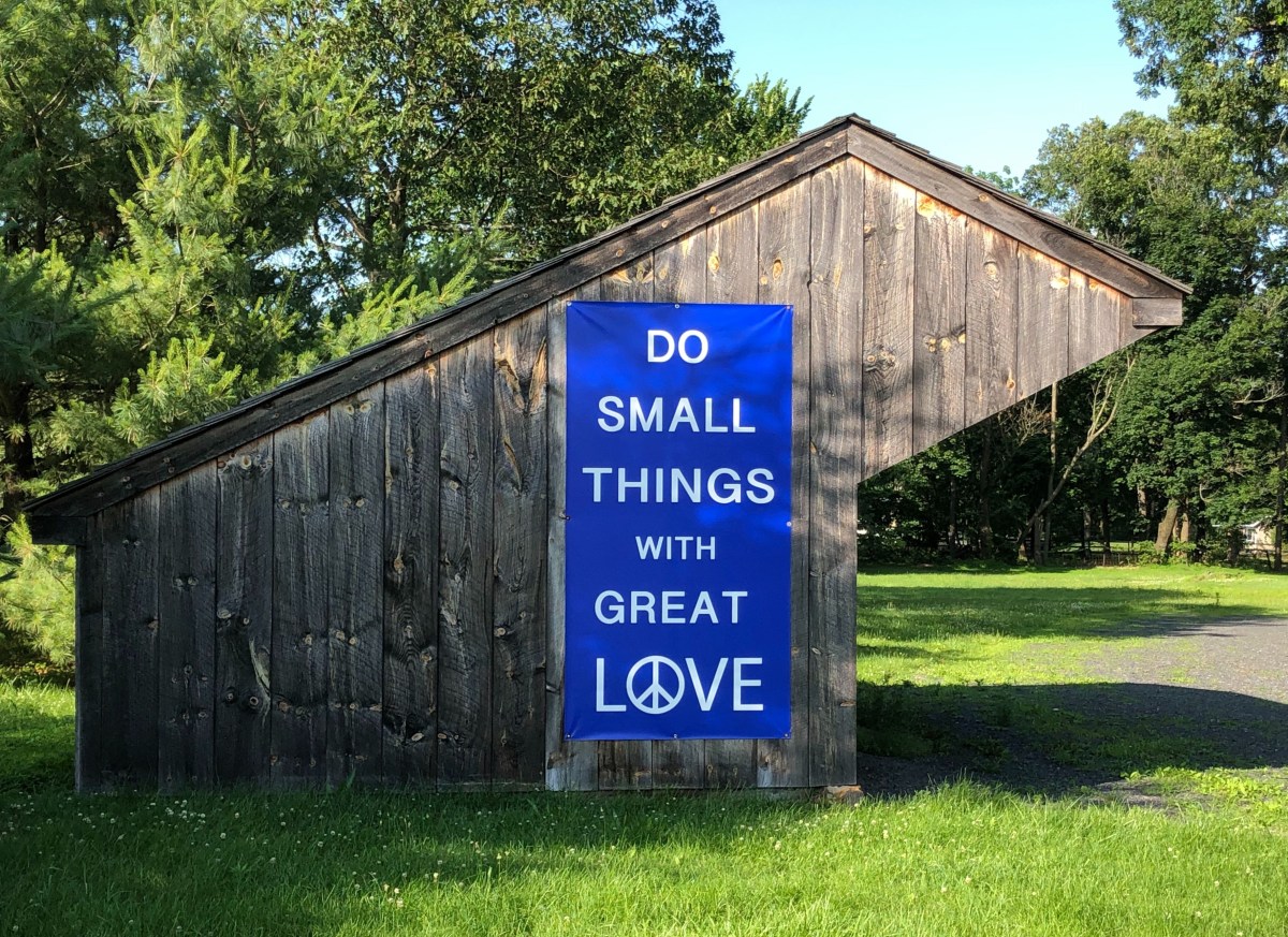 Photo shows wooden structure on meetinghouse grounds. One side of the structure features a large blue sign that says, in white writing, “Do small things with great love.” A peace sign is the “O” in “love.” Grass and trees appear in the foreground and background.